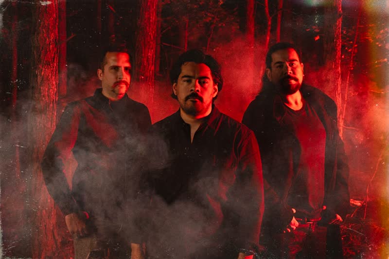 Tres músicos caminando por un bosque rojo frente a una capilla