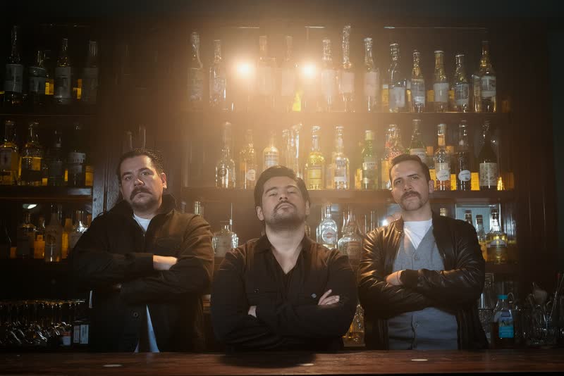 Trío de músicos con los brazos cruzados frente a una barra iluminada