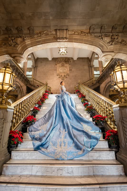 Quinceañera con vestido azul en una escalinata de palacio con flores rojas