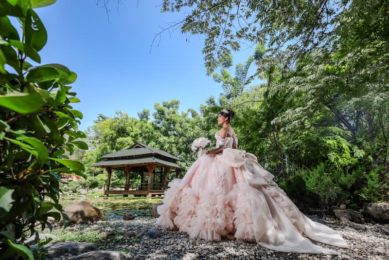 Quinceañera con vestido durazno en un jardín con un pabellón al fondo